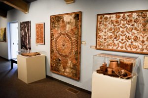 Another wall of the Wigwaazii Exhibit featuring contemporary wall art and historic baskets