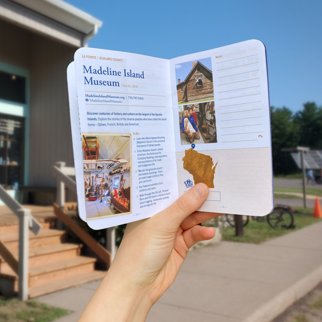 A hand holds up a passport book turned to the Madeline Island Museum page.